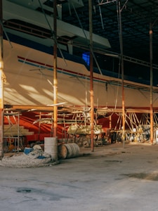 A large boat is situated inside a dimly lit, spacious dry dock or shipyard. Scaffolding surrounds the structure, indicating an ongoing construction or repair process. The foreground features various equipment such as ropes, barrels, and wooden planks scattered on a concrete floor.
