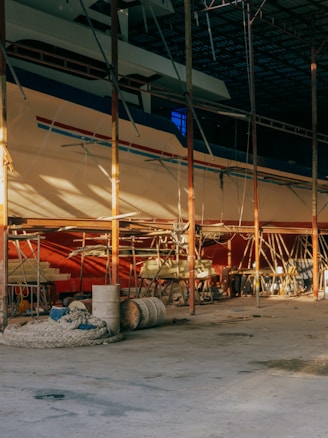 A large boat is situated inside a dimly lit, spacious dry dock or shipyard. Scaffolding surrounds the structure, indicating an ongoing construction or repair process. The foreground features various equipment such as ropes, barrels, and wooden planks scattered on a concrete floor.