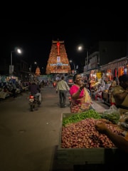 A bustling night market scene with people engaging in various activities. In the foreground, a vendor is selling green chilies and red onions. There is a woman in a pink and yellow saree, seemingly gesturing or talking. Behind, more vendors are selling goods under bright artificial lights. In the background, a brightly illuminated temple tower dominates the scene, creating a festive and vibrant atmosphere.