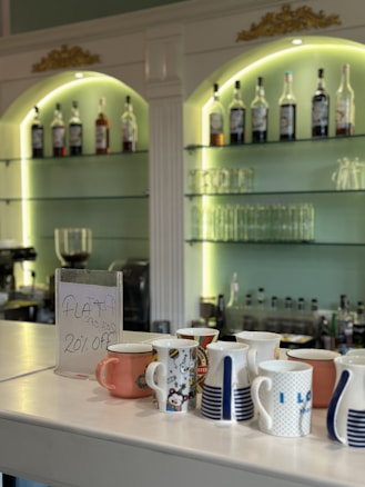A selection of colorful mugs is displayed on a countertop in front of a bar area. The sign near the mugs advertises a flat 20% off discount. Behind the counter, shelves are stocked with various bottles and glassware, illuminated by greenish lighting. The bar area features ornate decorations with gold accents at the top.