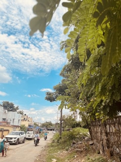 A rural road flanked by greenery, with a stone wall on one side and several people walking and riding motorcycles. The sky is mostly clear with a few white clouds, and the buildings in the background have a rustic appearance.