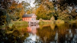 A small, charming brick house with a decorative white porch sits peacefully by the edge of a calm lake, surrounded by lush green and autumnal trees. The reflections of the house and trees create a mirror-like effect on the water's surface. The scene is framed by thick foliage in the foreground, providing a sense of seclusion and tranquility.