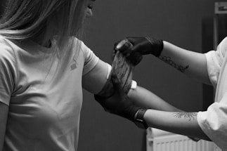 A person wearing a t-shirt is receiving an arm wrap or bandage from another individual whose arms have tattoos and is wearing black gloves. The setting is indoors, with a radiator visible in the background.