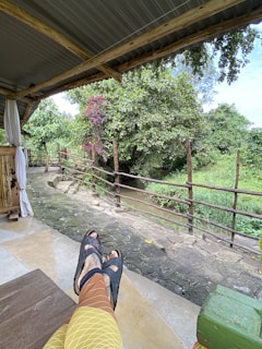 A person is relaxing by sitting with their feet up on a wooden bench under a canopy. The scene overlooks a serene natural setting featuring lush greenery. A small, tranquil stream can be seen in the background, bordered by a wooden fence. The area is shaded by a large leafy tree with some branches bearing reddish leaves.