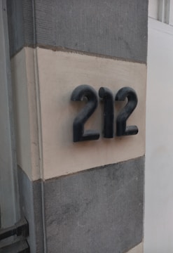Metallic numbers 212 are mounted on a light-colored wall, framed by darker vertical stripes. The numbers have a three-dimensional, rounded design.