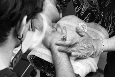 A tattoo artist is working on a person's hand, applying a tattoo design. The setting appears to be a studio, with the client's arm resting on a padded surface. The tattoo artist wears gloves and is in the process of tattooing a design that seems to feature a fish or aquatic creature.