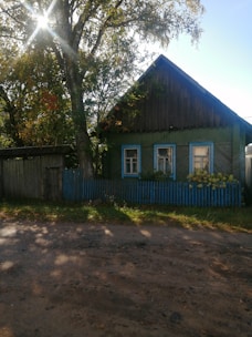 A rustic wooden house with a green facade and blue window frames is partly obscured by large trees. The sunlight filters through the leaves, creating a gentle dappled effect on the dirt road in front. A small picket fence, also painted blue, surrounds the house, and some yellow flowers add a touch of color near the windows.