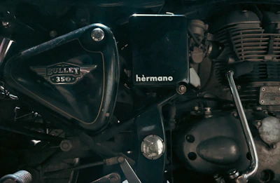 A close-up view of the side of a motorcycle engine, showcasing the intricate mechanical parts and branding details such as 'Bullet 350' and 'hermano'. The image focuses on the metallic and polished components, highlighting the craftsmanship of the motorcycle.