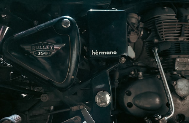A close-up view of the side of a motorcycle engine, showcasing the intricate mechanical parts and branding details such as 'Bullet 350' and 'hermano'. The image focuses on the metallic and polished components, highlighting the craftsmanship of the motorcycle.