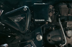 A close-up view of the side of a motorcycle engine, showcasing the intricate mechanical parts and branding details such as 'Bullet 350' and 'hermano'. The image focuses on the metallic and polished components, highlighting the craftsmanship of the motorcycle.