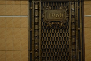 A metallic ornate grill with decorative patterns and the number 1960 prominently displayed in the center. The grill is framed with intricate designs and the surrounding wall is tiled with a neutral beige color.
