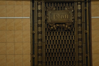 A metallic ornate grill with decorative patterns and the number 1960 prominently displayed in the center. The grill is framed with intricate designs and the surrounding wall is tiled with a neutral beige color.