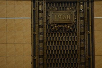 A metallic ornate grill with decorative patterns and the number 1960 prominently displayed in the center. The grill is framed with intricate designs and the surrounding wall is tiled with a neutral beige color.