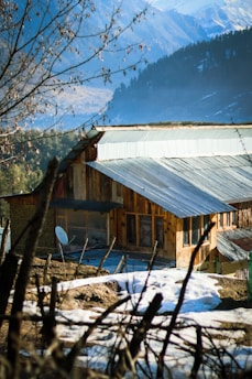 A rustic wooden house with a metal roof is situated in a mountainous area. The building is surrounded by snow-covered ground and bare trees. The background features a range of mountains partly covered in snow, against a clear blue sky.