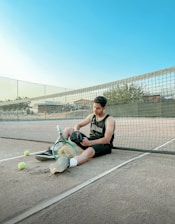 A person is seated on a tennis court, leaning against a net. They are dressed in athletic wear and holding a cap. Near them, there are tennis balls and a racket lying on the ground. The sky is clear and blue, suggesting pleasant weather.