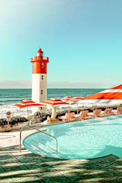 A brightly lit coastal scene featuring a red and white lighthouse situated by the sea. A swimming pool in the foreground is surrounded by red and white striped umbrellas and sun loungers. The setting exudes a summery, relaxing atmosphere with clear blue skies and calm ocean waters.