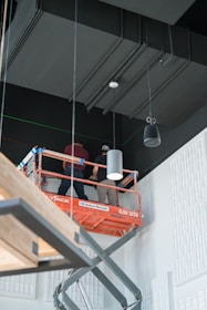Two workers are standing on an orange scissor lift inside a building. They are working near the ceiling, which is painted black with square acoustic panels on the walls. The lift is labeled with a rental company name. The environment appears to be industrial or under construction.