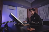 A person sits at a desk in a modern studio, working on a computer. The room features soundproof panels on the walls and ambient lighting.