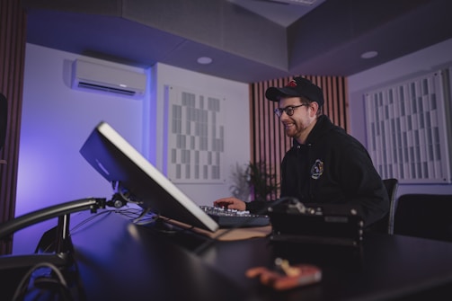 A person sits at a desk in a modern studio, working on a computer. The room features soundproof panels on the walls and ambient lighting.