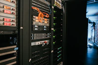 A data center with rows of servers in racks, featuring various network and power connections. Orange and black cables are visible, alongside multiple server units with indicator lights. The environment appears organized with subdued lighting.