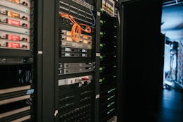 A data center with rows of servers in racks, featuring various network and power connections. Orange and black cables are visible, alongside multiple server units with indicator lights. The environment appears organized with subdued lighting.