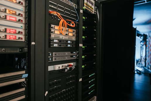 A data center with rows of servers in racks, featuring various network and power connections. Orange and black cables are visible, alongside multiple server units with indicator lights. The environment appears organized with subdued lighting.