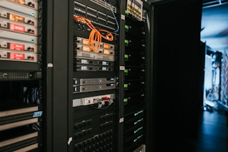 A data center with rows of servers in racks, featuring various network and power connections. Orange and black cables are visible, alongside multiple server units with indicator lights. The environment appears organized with subdued lighting.