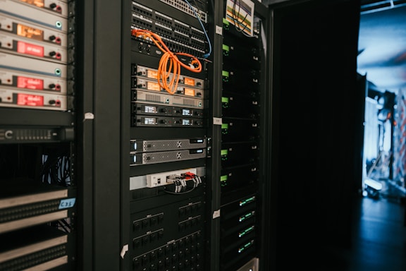 A data center with rows of servers in racks, featuring various network and power connections. Orange and black cables are visible, alongside multiple server units with indicator lights. The environment appears organized with subdued lighting.