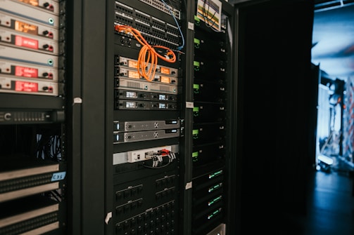 A data center with rows of servers in racks, featuring various network and power connections. Orange and black cables are visible, alongside multiple server units with indicator lights. The environment appears organized with subdued lighting.