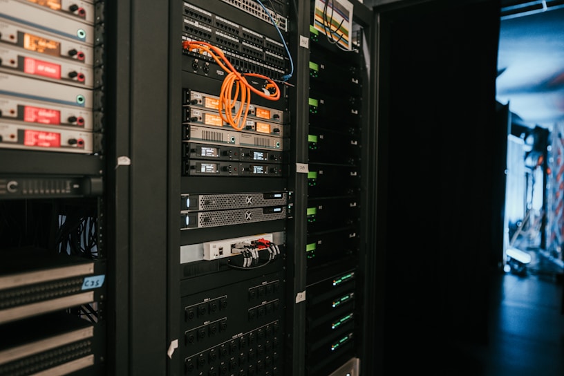 A data center with rows of servers in racks, featuring various network and power connections. Orange and black cables are visible, alongside multiple server units with indicator lights. The environment appears organized with subdued lighting.