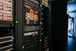 A data center with rows of servers in racks, featuring various network and power connections. Orange and black cables are visible, alongside multiple server units with indicator lights. The environment appears organized with subdued lighting.