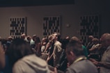 A diverse group of people stand closely together in a dimly lit room, possibly engaged in a contemplative or solemn activity. Acoustic panels are visible on the walls, and everyone appears focused, contributing to a quiet and introspective atmosphere.