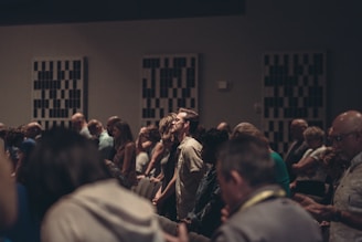 A diverse group of people stand closely together in a dimly lit room, possibly engaged in a contemplative or solemn activity. Acoustic panels are visible on the walls, and everyone appears focused, contributing to a quiet and introspective atmosphere.