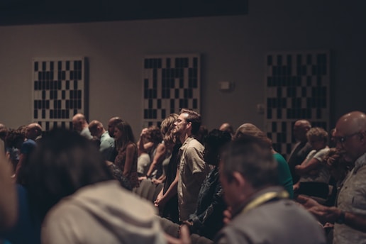A diverse group of people stand closely together in a dimly lit room, possibly engaged in a contemplative or solemn activity. Acoustic panels are visible on the walls, and everyone appears focused, contributing to a quiet and introspective atmosphere.