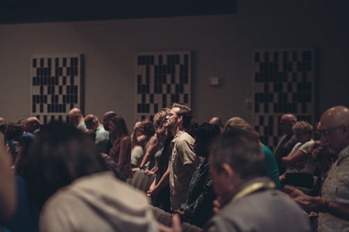 A diverse group of people stand closely together in a dimly lit room, possibly engaged in a contemplative or solemn activity. Acoustic panels are visible on the walls, and everyone appears focused, contributing to a quiet and introspective atmosphere.