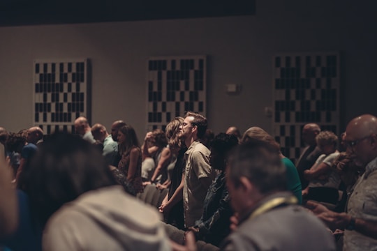 A diverse group of people stand closely together in a dimly lit room, possibly engaged in a contemplative or solemn activity. Acoustic panels are visible on the walls, and everyone appears focused, contributing to a quiet and introspective atmosphere.