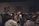 A diverse group of people stand closely together in a dimly lit room, possibly engaged in a contemplative or solemn activity. Acoustic panels are visible on the walls, and everyone appears focused, contributing to a quiet and introspective atmosphere.