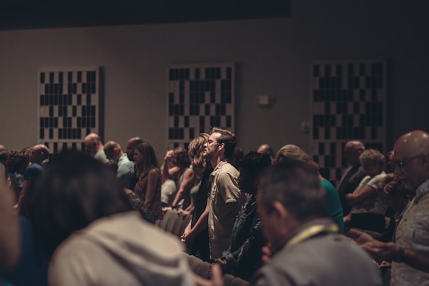 A diverse group of people stand closely together in a dimly lit room, possibly engaged in a contemplative or solemn activity. Acoustic panels are visible on the walls, and everyone appears focused, contributing to a quiet and introspective atmosphere.