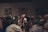A diverse group of people stand closely together in a dimly lit room, possibly engaged in a contemplative or solemn activity. Acoustic panels are visible on the walls, and everyone appears focused, contributing to a quiet and introspective atmosphere.