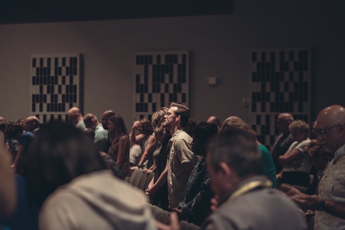 A diverse group of people stand closely together in a dimly lit room, possibly engaged in a contemplative or solemn activity. Acoustic panels are visible on the walls, and everyone appears focused, contributing to a quiet and introspective atmosphere.