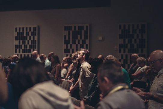 A diverse group of people stand closely together in a dimly lit room, possibly engaged in a contemplative or solemn activity. Acoustic panels are visible on the walls, and everyone appears focused, contributing to a quiet and introspective atmosphere.