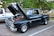 A classic black pickup truck is parked in an outdoor setting with its hood raised, revealing a detailed and clean engine. The truck features polished chrome accents and shiny wheels. Surrounding the truck are green trees, pavement, and a background that includes another white truck and various park elements.