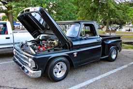 A classic black pickup truck is parked in an outdoor setting with its hood raised, revealing a detailed and clean engine. The truck features polished chrome accents and shiny wheels. Surrounding the truck are green trees, pavement, and a background that includes another white truck and various park elements.