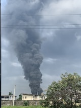 A large column of thick black smoke rises high into the overcast sky above a building. The scene includes a residential structure, utility lines, and a tree in the foreground with a partly cloudy sky.