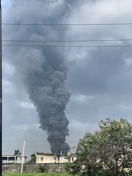 A large column of thick black smoke rises high into the overcast sky above a building. The scene includes a residential structure, utility lines, and a tree in the foreground with a partly cloudy sky.