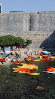 A group of colorful kayaks, mostly in red and yellow, are lined up near a pebbly shore. Several people are gathered around the area, some preparing or tending to the kayaks. A massive stone fortification forms the backdrop, with patches of greenery on one side.
