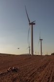 A series of large wind turbines are spread out across a grassy field under a clear sky. In the foreground, a person lies on the ground, possibly resting or observing. A drone is hovering in the air near the turbines.