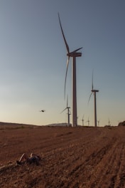 A series of large wind turbines are spread out across a grassy field under a clear sky. In the foreground, a person lies on the ground, possibly resting or observing. A drone is hovering in the air near the turbines.