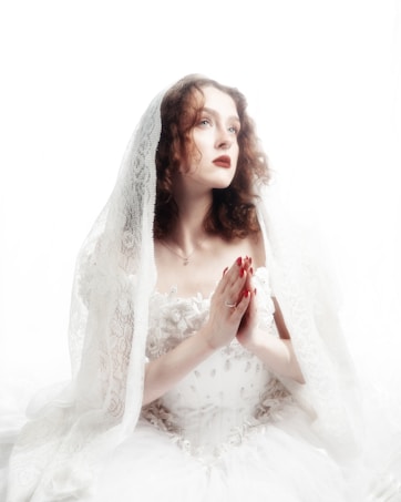 A woman with curly hair sits with hands clasped in prayer. She wears a white lace veil and a detailed white gown adorned with floral motifs, set against a white background. Her expression appears serene and contemplative, with a focused upward gaze.
