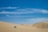 A vast, sandy desert landscape under a clear, blue sky. A four-wheel-drive vehicle is seen navigating the dunes, leaving tracks in the sand behind it. The dunes are gently curving and stretch towards the horizon.